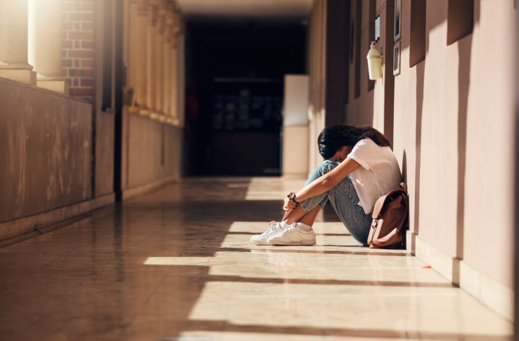 A teenage girl sits on the floor in a high school hallway with her backpack next to her. She's sitting with her legs crossed, arms over her legs, and her head down on her knees