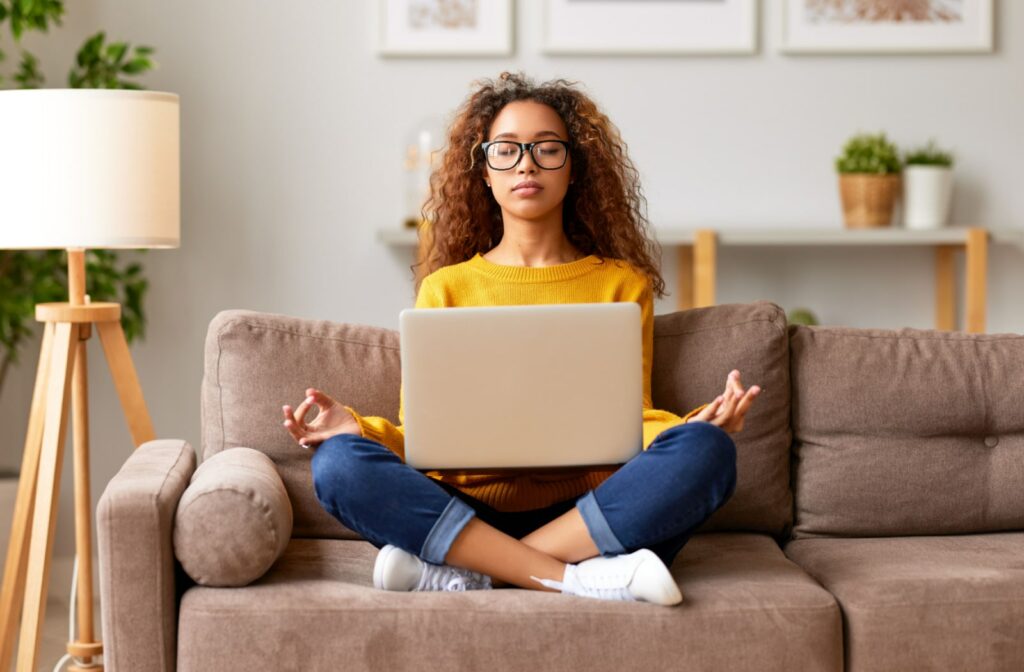 A teenage girl sitting with her legs crossed on the couch, and her laptop is also sitting on her lap. She is trying to meditate