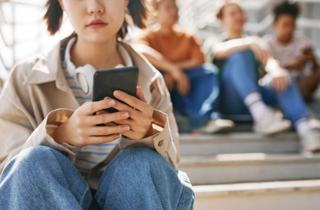 A youth sitting on a stairs outside in front of a group of other youth texting a mental health support line, like ConnecTeen.