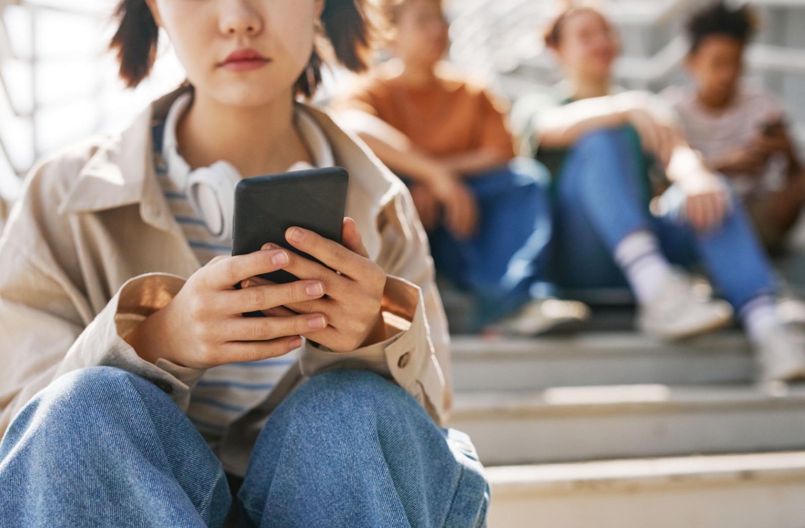 A youth sitting on a stairs outside in front of a group of other youth texting a mental health support line, like ConnecTeen.